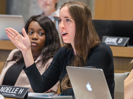A student speaks with her hand raised at the Kellogg Climate Conference, as two students watch her in the background. 