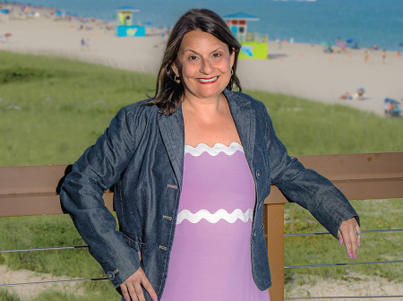 Maria Atanasov poses on a boardwalk with her arms propped up on a wooden railing in front of a sunny beach in Florida.