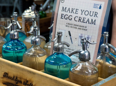 A wooden crate holds blue and tan bottles with ingredients to make an egg cream.