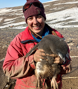 Parker Levinson is wearing a red parka and a maroon knit hat, and she is holding a penguin by its midriff. Icy Antarctica background.